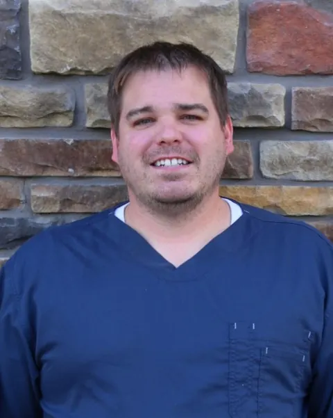 Dr. Jerad Hintz, DDS, smiling in navy scrubs in front of a stone wall